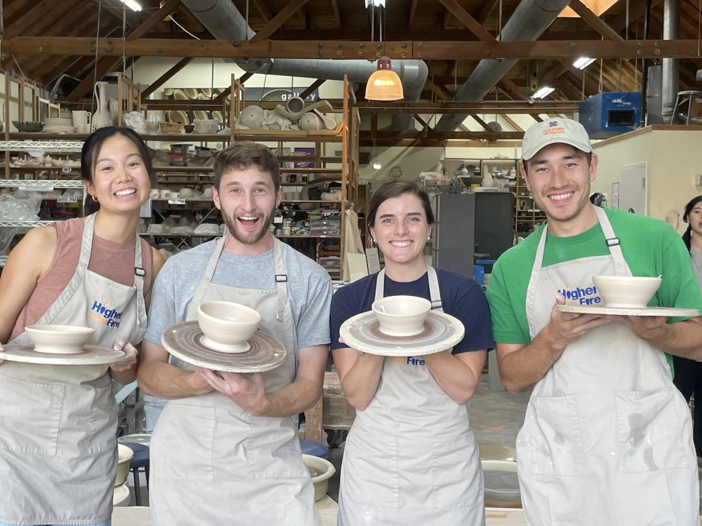 Four people wearing tan Higher Fire aprons smiling and holding freshly thrown pots.